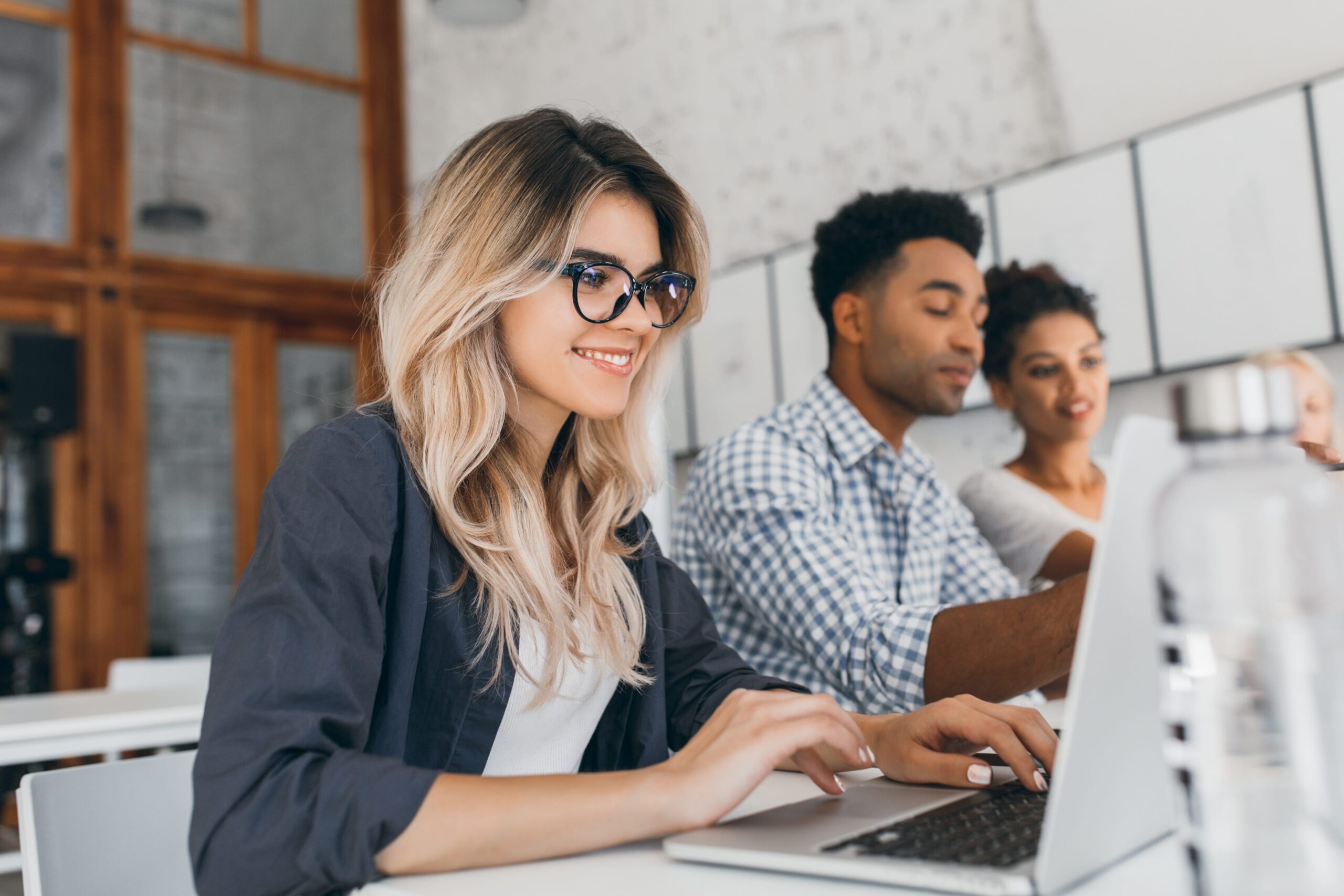 beautiful-curly-female-freelancer-with-cute-manicure-using-laptop-smiling-indoor-portrait-blonde-secretary-sitting-beside-african-coworker-blue-shirt-min