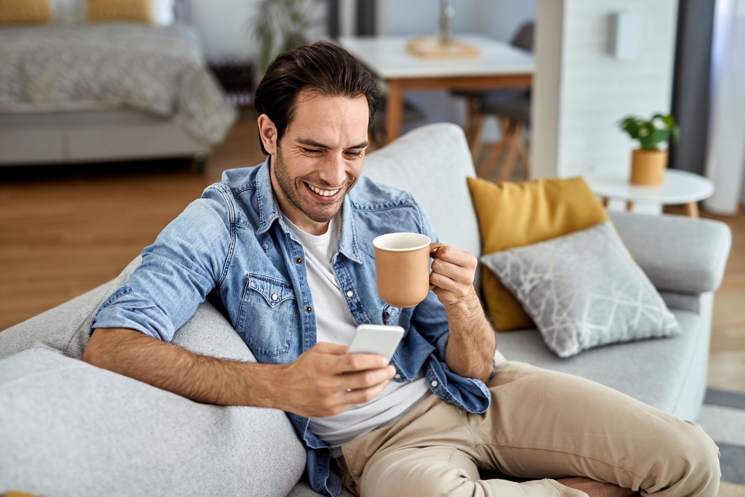 young-happy-man-texting-smart-phone-while-relaxing-sofa-drinking-coffee-min